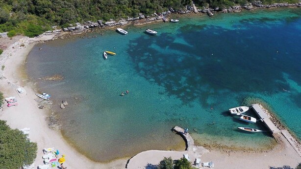 Vista aérea de una playa con aguas turquesas cristalinas, embarcaciones y un pequeño muelle.