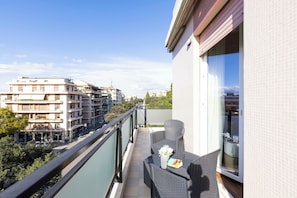 A balcony with a wicker table and chairs, a glass door leading to a room, and a view of surrounding buildings.