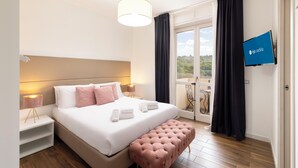 A hotel room with a bed, bedside lamps, a TV, a balcony with curtains, and a wooden floor. - Mapi's rooms (Cagliari)