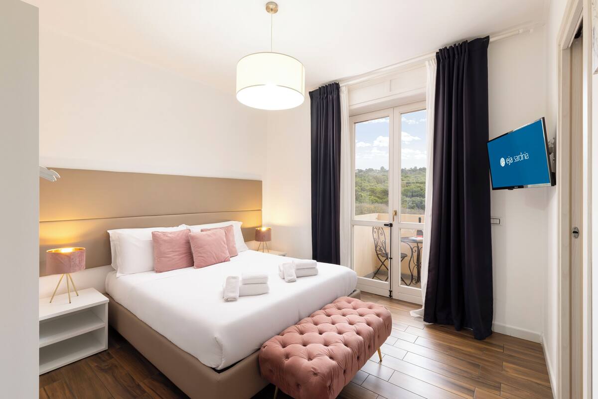 Una habitación de hotel con cama, lámparas de noche, televisión, balcón con cortinas y suelo de madera.