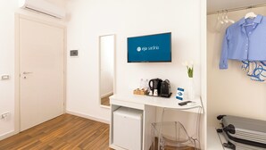 A hotel room with a white desk, a television displaying 'eja sardinia', a small refrigerator, a clear chair, a hanging coat, and a suitcase. - Mapi's rooms (Cagliari)
