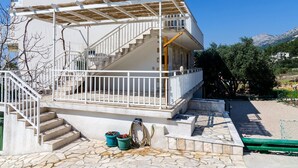 Un edificio de dos plantas con fachada blanca, un espacio al aire libre cubierto con una pérgola y un acceso empedrado con piedra.