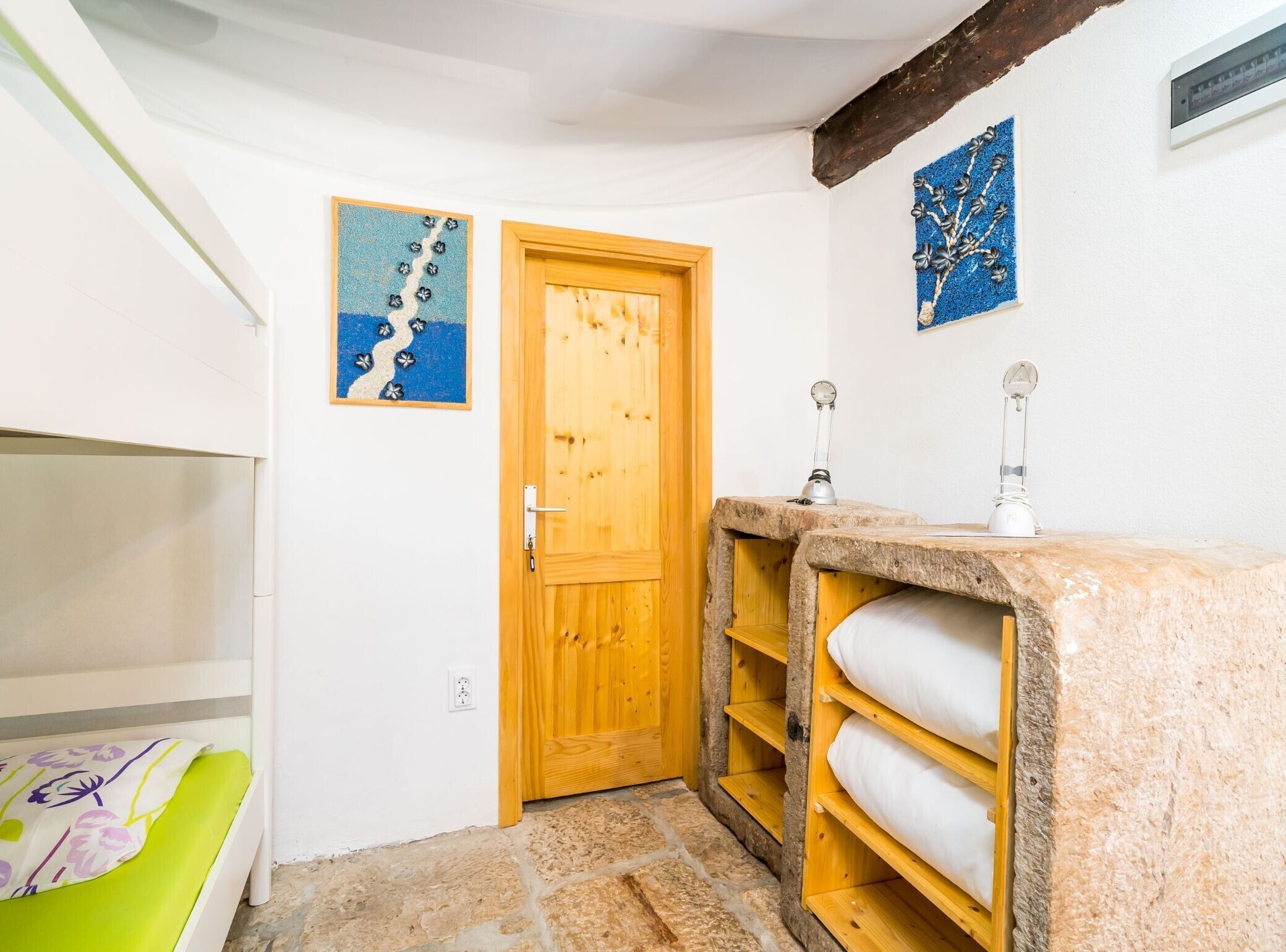 A small, cozy room with a wooden door, a stone floor, a bed with a green and white cover, and a stone bench.