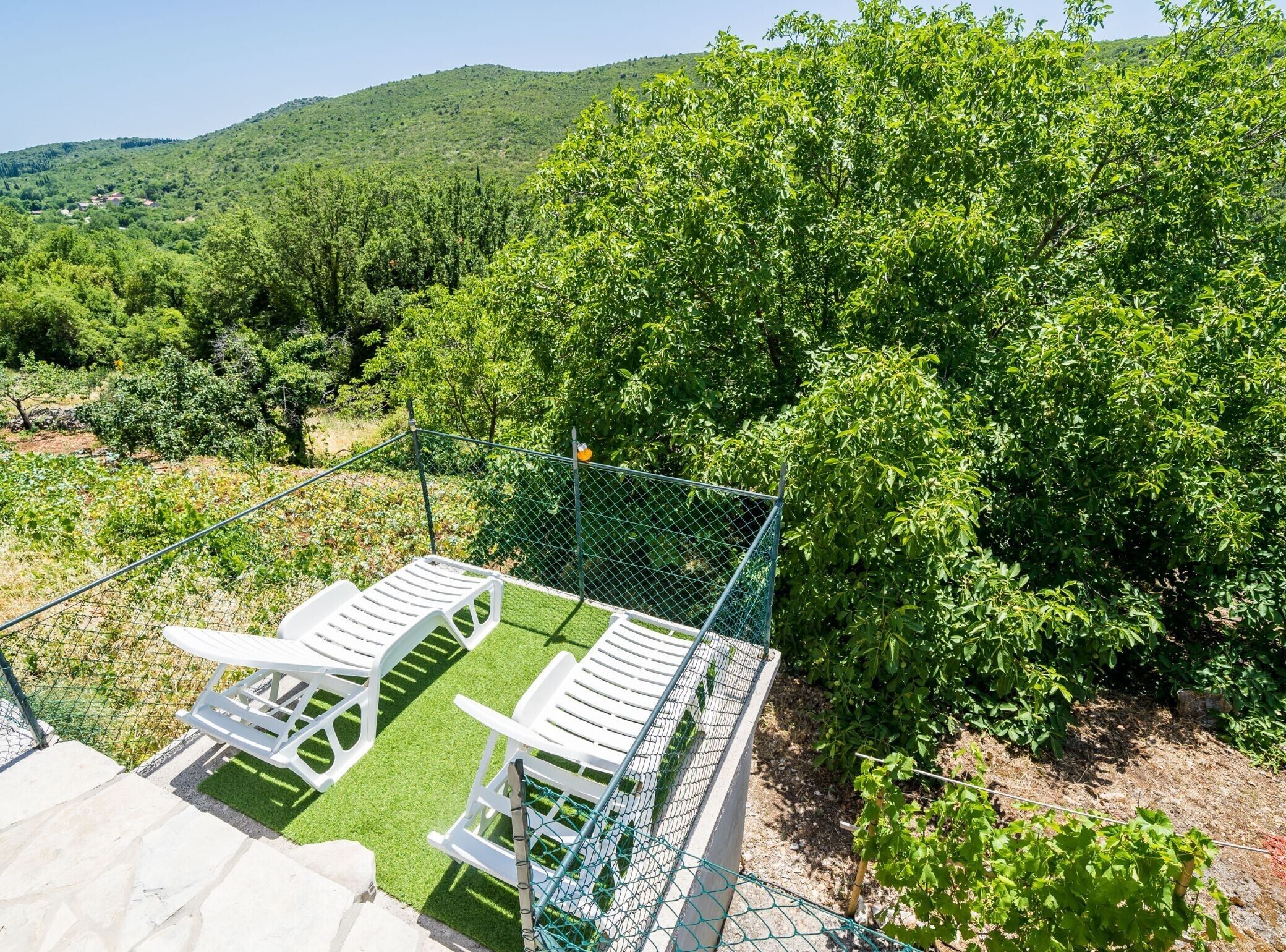 Un área exterior cercada con sillas de plástico blanco y una mesa pequeña, rodeada de abundante vegetación y un paisaje montañoso al fondo.