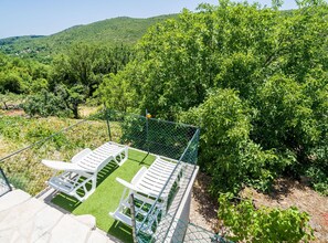 Un área exterior cercada con sillas de plástico blanco y una mesa pequeña, rodeada de abundante vegetación y un paisaje montañoso al fondo.
