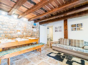 Una habitación rústica con paredes de piedra, vigas de madera, suelo de piedra, una mesa de madera con platos, un banco, un sofá y una alfombra estampada.