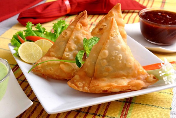 A plate of samosas with a side of chutney and a small bowl of sauce, garnished with mint leaves and a slice of lemon.