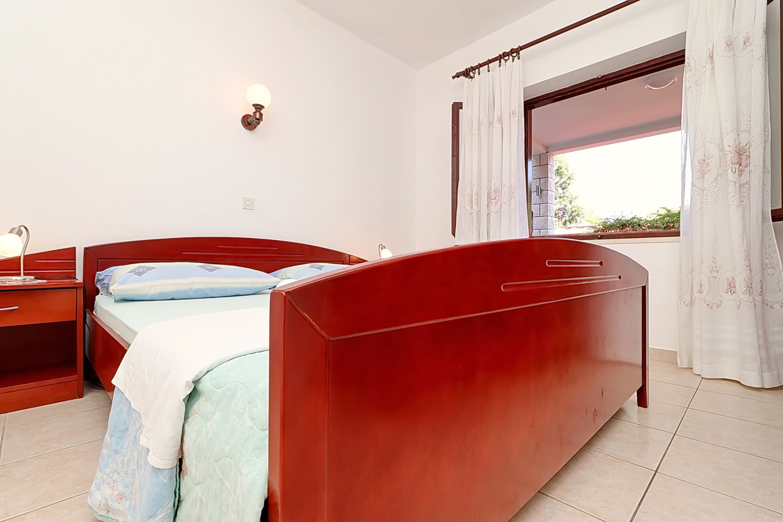 Une chambre à coucher avec un lit rouge, une table de chevet, une lampe et une fenêtre avec des rideaux.