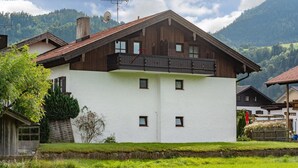 Une maison en bois à deux étages, dotée d’un balcon, entourée de verdure et de montagnes.