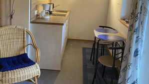 Une cuisine compacte équipée d’une machine à café, d’une petite table à manger avec des chaises et d’un fauteuil en osier avec un coussin bleu.