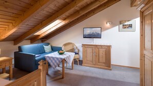 Une pièce chaleureuse avec un plafond en pente, des poutres en bois, un canapé bleu, une petite table avec un saladier, une chaise en osier, un meuble en bois et un téléviseur fixé au mur.