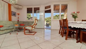 Un salon avec un canapé, une table et des chaises, un lustre et une vue sur l’extérieur, visible à travers une porte.