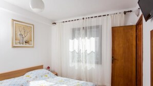 A bedroom with a bed, a wooden door, a window with sheer curtains, and a painting on the wall.