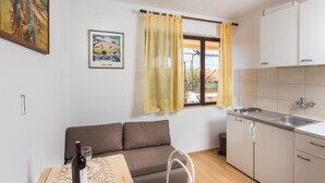 A small, well-lit room with a sofa, a dining table with a bottle and glass, a window with yellow curtains, and a kitchenette with a sink and cabinets.