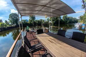 A covered deck with wicker furniture and a table, overlooking a calm lake with boats docked. - Prague Bay Houseboats (Prague)