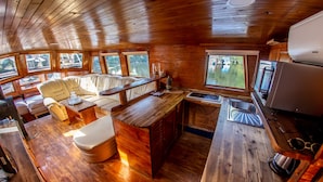 A compact cabin interior with a wooden floor, a sofa, a dining table, a kitchen area with a sink and stove, and large windows offering views of the outdoors. - Prague Bay Houseboats (Prague)