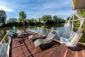 A deck with lounge chairs and a hammock, overlooking a calm river with trees and buildings in the background. - Prague Bay Houseboats (Prague)