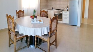 A dining area with a round table, chairs, and a fruit bowl. A kitchen with white cabinets and appliances is visible in the background.