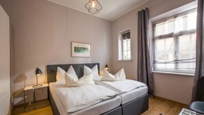 Cama doble con ropa de cama blanca, mesita de noche con lámpara, un cuadro en la pared y una ventana con cortinas.