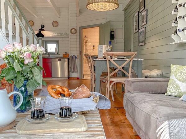 A cozy living room with a grey sofa, a dining table with pretzels and bread, a vase of flowers, and a kitchen in the background.