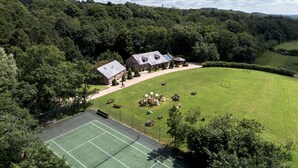 A large house with a tennis court, surrounded by a lush green landscape and dense forest.