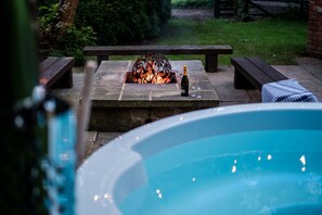 Un foyer en pierre avec une bouteille et des verres, entouré de bancs en bois, dans un jardin avec une piscine.