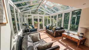 Ein Wintergarten mit Glasdach, ausgestattet mit Ledersofas und einem Holztisch, umgeben von großen Fenstern.