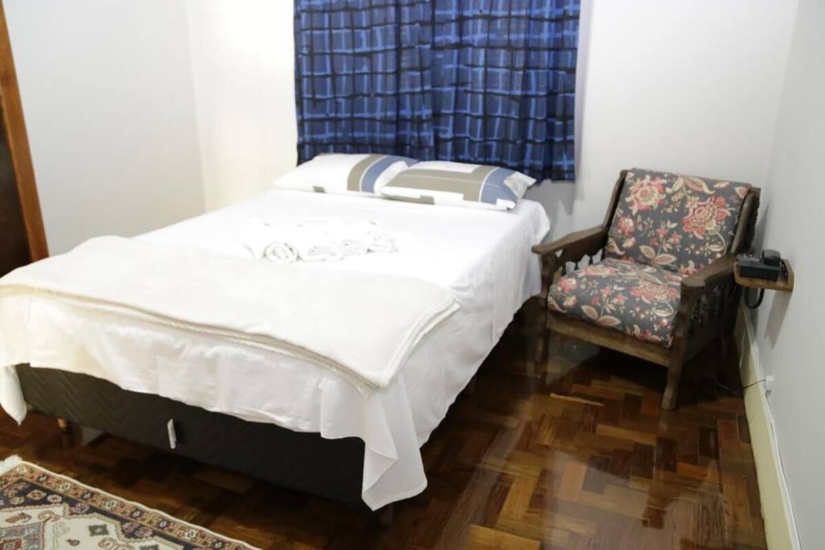 Ein Schlafzimmer mit einem Bett, einem Sessel, einem gemusterten Teppich und einem Fenster mit blauen Vorhängen.