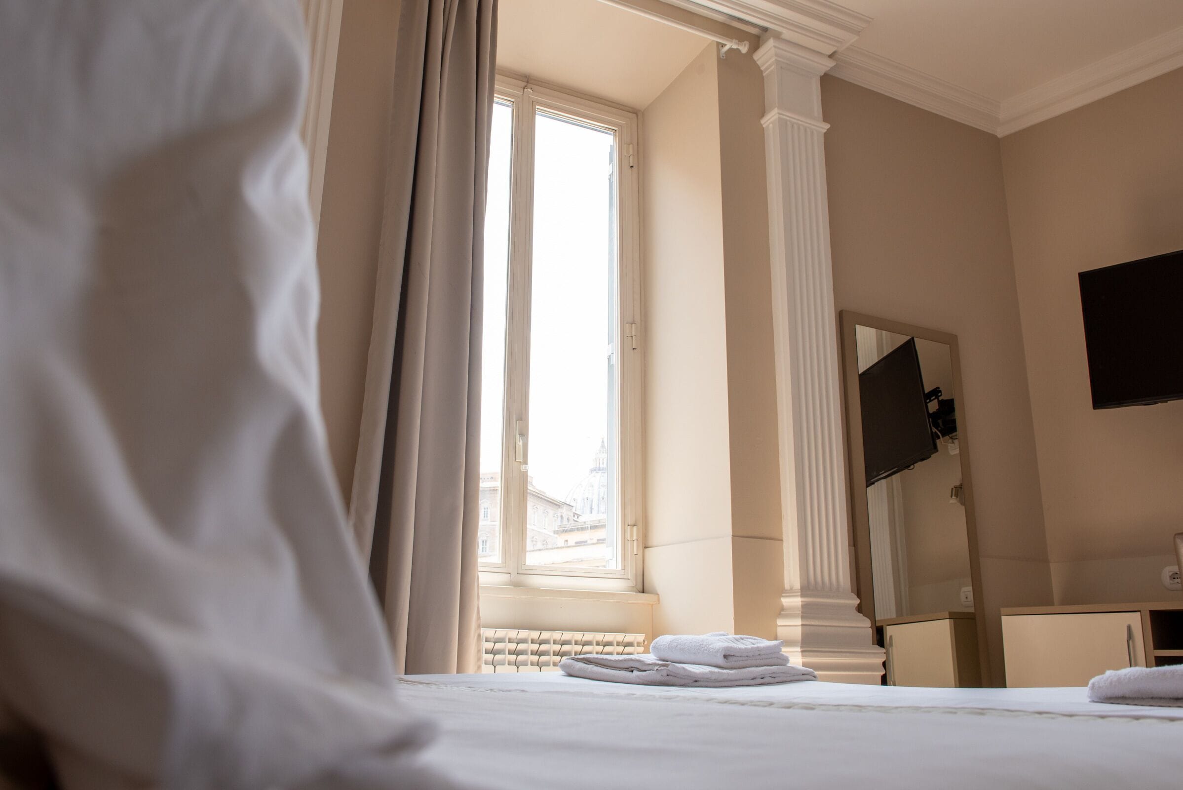 Une chambre d’hôtel avec un lit, une fenêtre avec des rideaux, un radiateur, un miroir et un téléviseur.