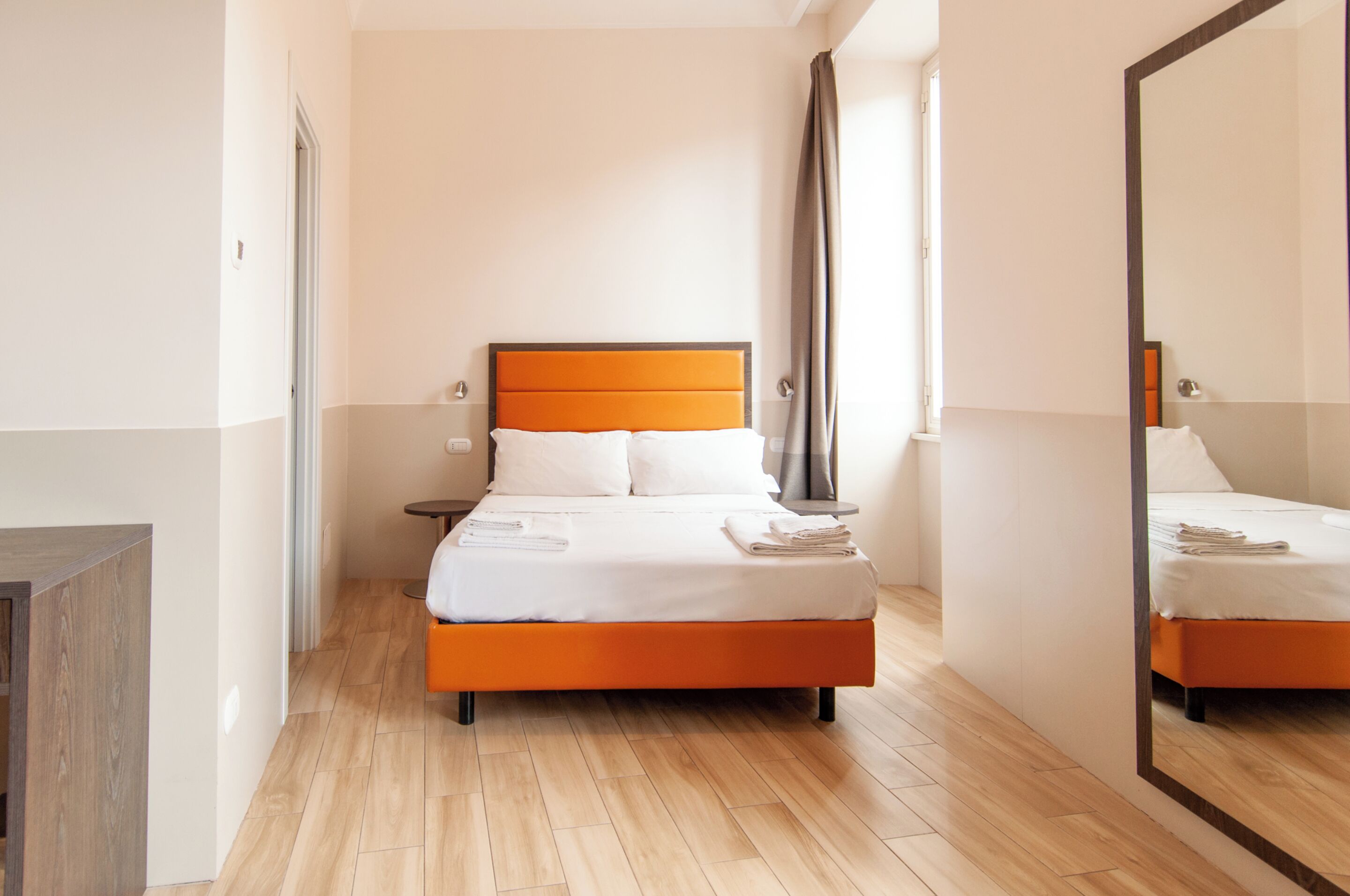 A modern hotel room with a wooden floor, a bed with an orange headboard, white bedding, and a large mirror on one wall.