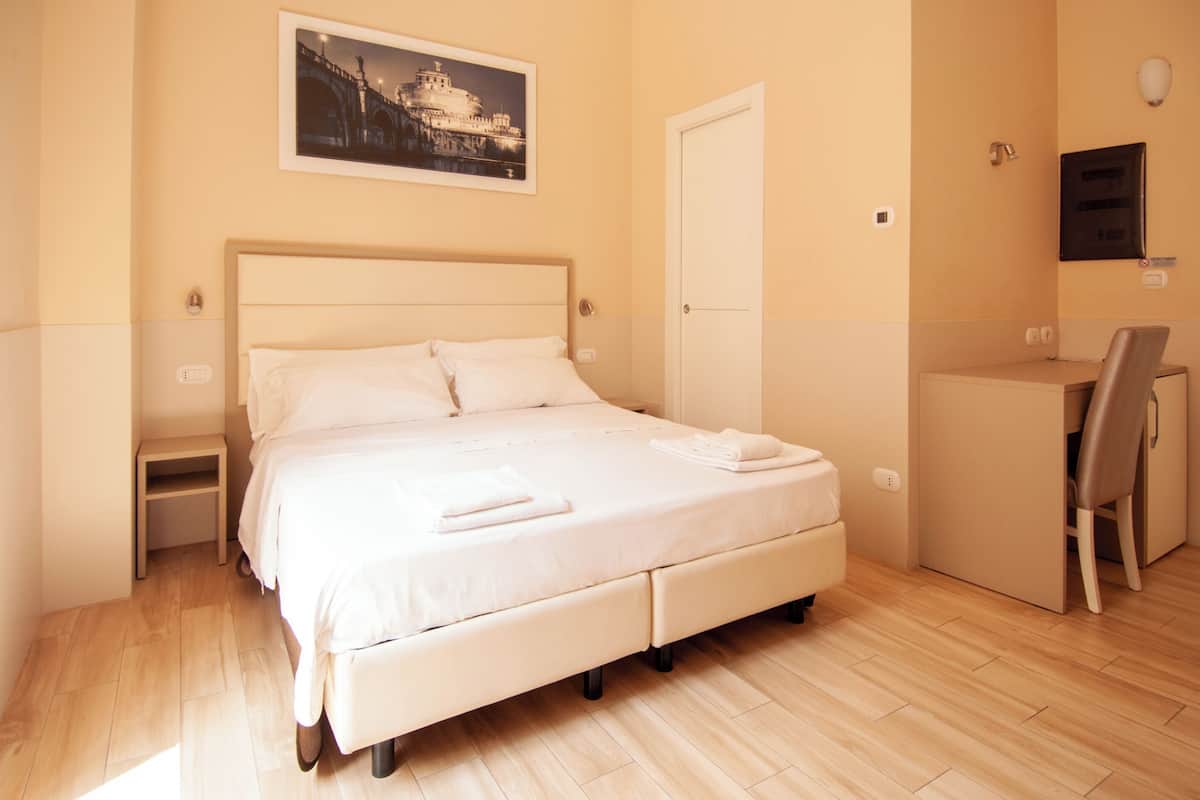 A neatly made bed with pillows and a folded towel, a bedside table, a desk with a chair, and a framed picture on the wall.