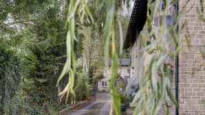 Ein schmaler gepflasterter Weg führt zu einem steinernen Gebäude mit traditioneller Gestaltung, umgeben von dichtem Grün und Bäumen.