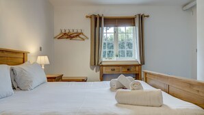 Ein ordentlich bezogenes Bett mit weißen Leinen, ein Nachttisch mit Lampe, ein Fenster mit Vorhängen und ein Holz-Kopfteil.