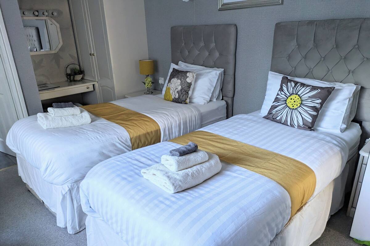 A hotel room with two beds, each with white bedding and a gold throw blanket. There is a nightstand with a lamp and a mirror above it.