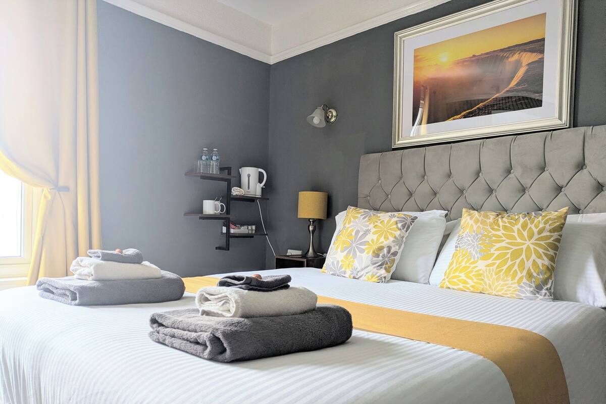 A hotel room with a neatly made bed, a bedside table with a lamp, a shelf with toiletries, and a framed picture on the wall.