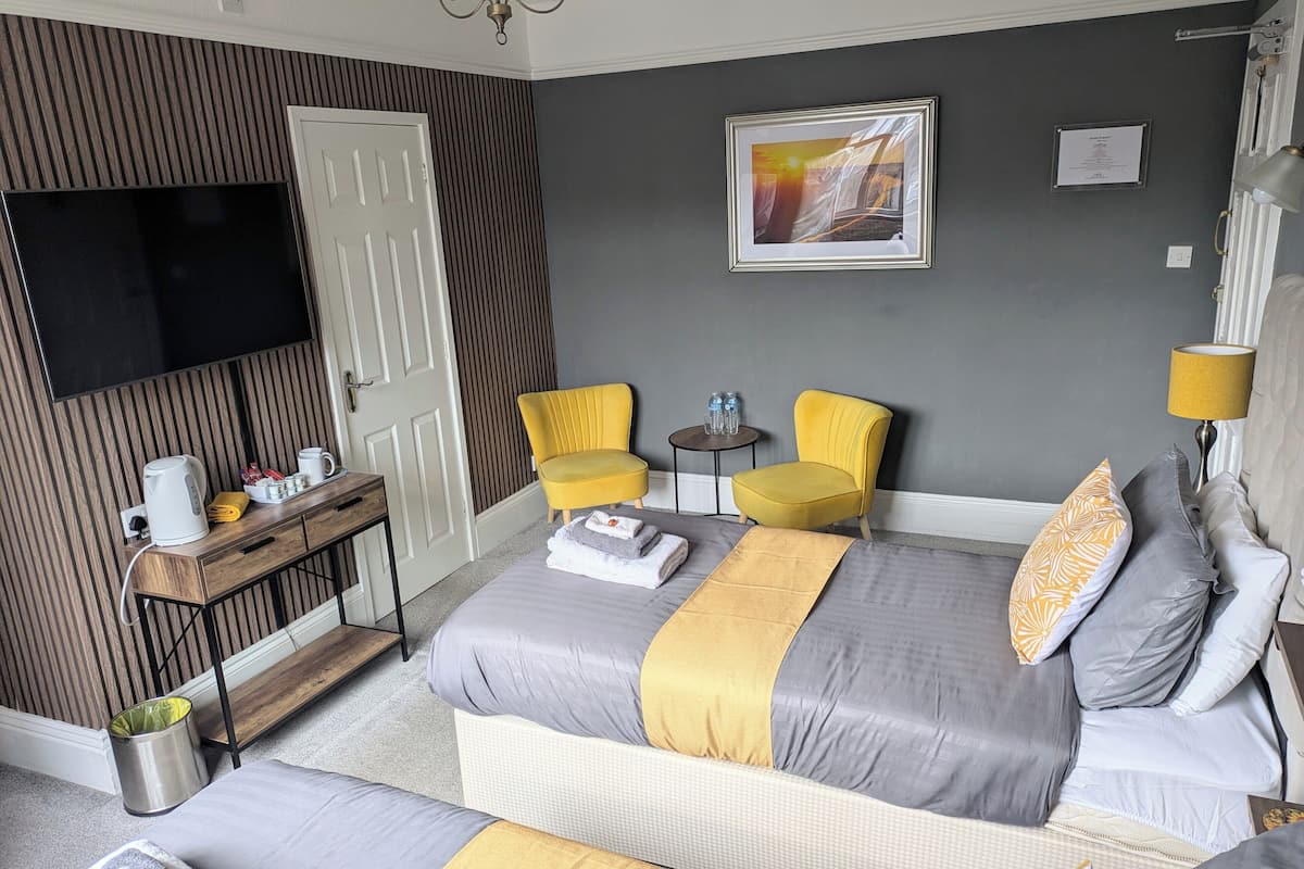 A hotel room with a bed, a TV, a small table with drinks, a chair, and a lamp.