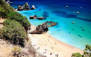 Une plage côtière aux eaux turquoise et limpides, parsemée de rochers et où l’on aperçoit quelques bateaux.