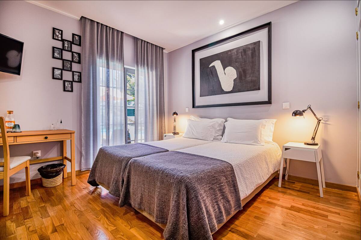 Een slaapkamer met een tweepersoonsbed, een bureau, een stoel, een bijzettafeltje, een lamp en een lijst met een kunstwerk aan de muur.