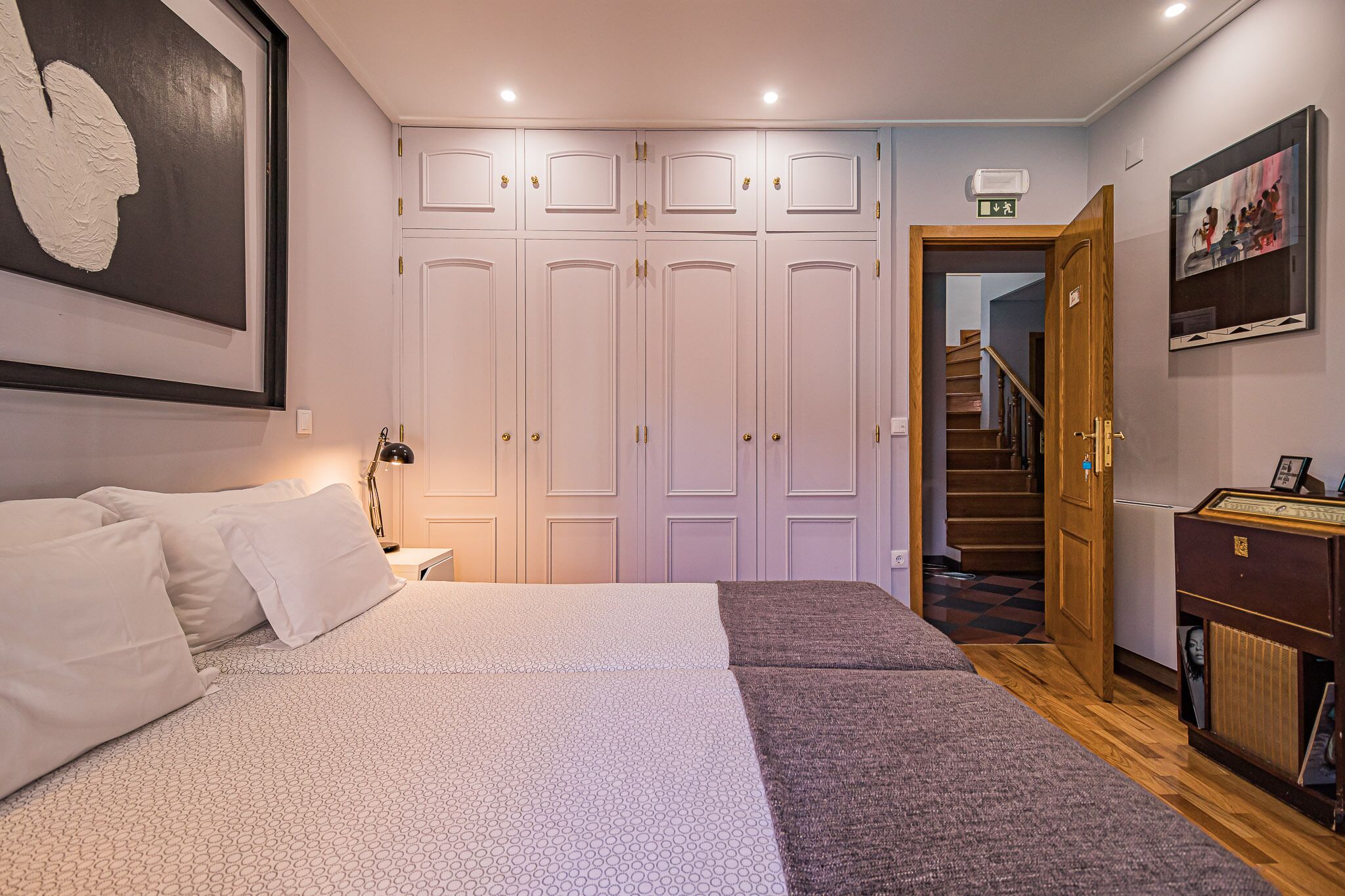 A bedroom with a large bed, a wardrobe, a desk with a lamp, and a staircase leading to another floor.