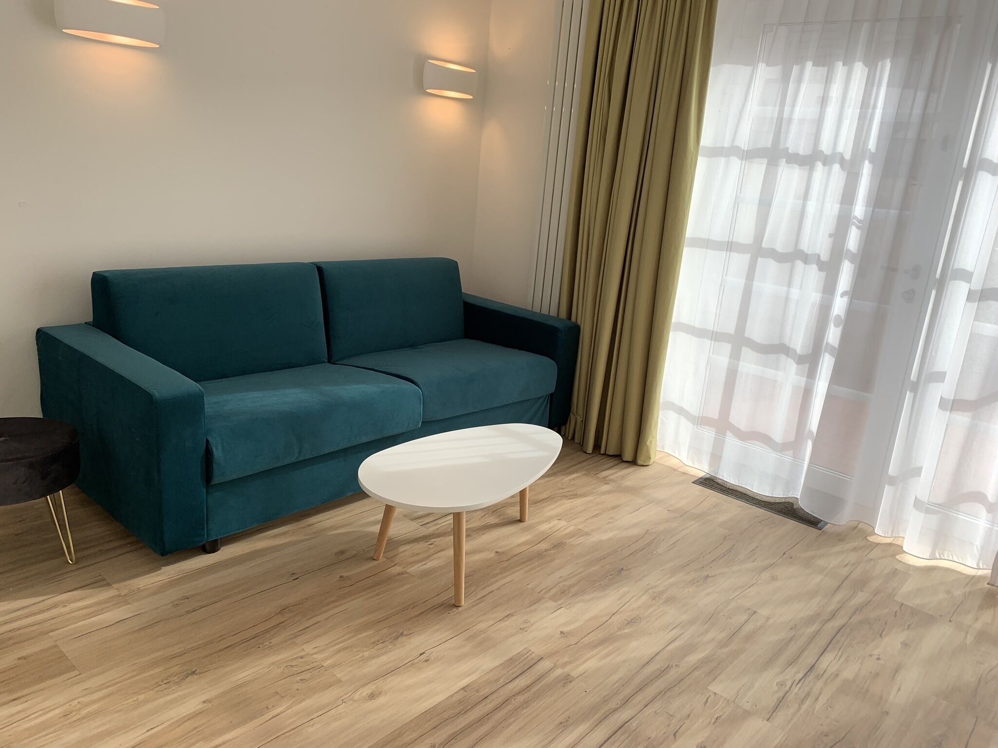 Um sofá azul-esverdeado, uma mesa de centro branca e um chão de madeira num quarto com janela e cortinas.