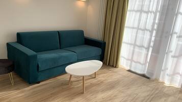 Um sofá azul-esverdeado, uma mesa de centro branca e um chão de madeira num quarto com janela e cortinas.