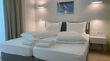 Una habitación de hotel moderna con una cama grande, ropa de cama blanca, cabecero con iluminación integrada, una mesita de noche pequeña y una obra de arte en la pared que representa veleros.