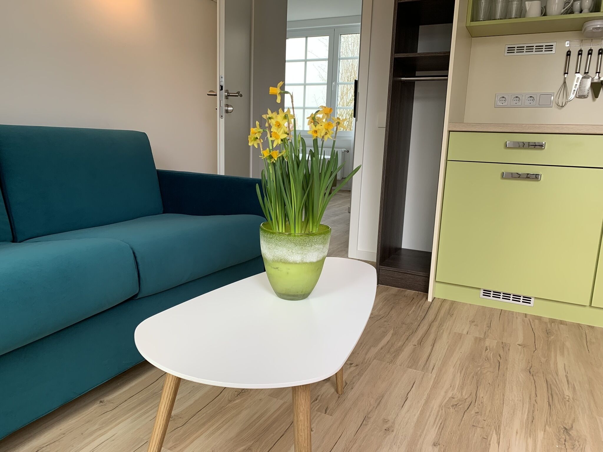 A room with a teal sofa, a white table with a green vase of daffodils, and a green kitchenette.