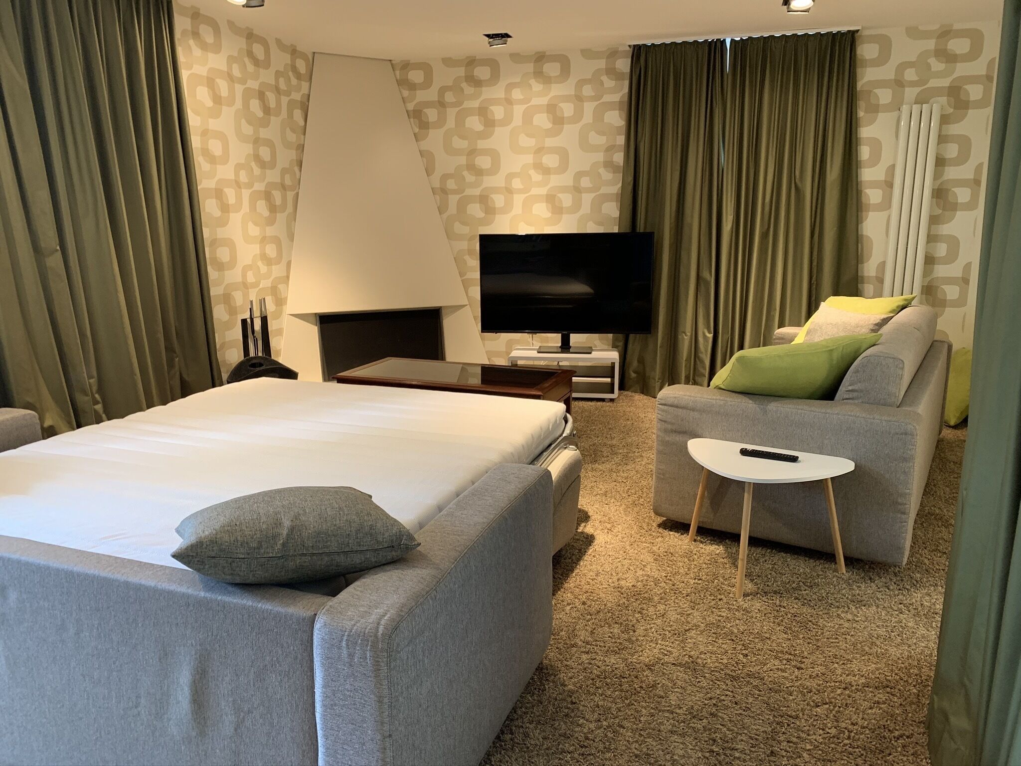 Habitació d'hotel amb un llit gran, un sofà gris, una taula petita blanca, una televisió i una làmpada.