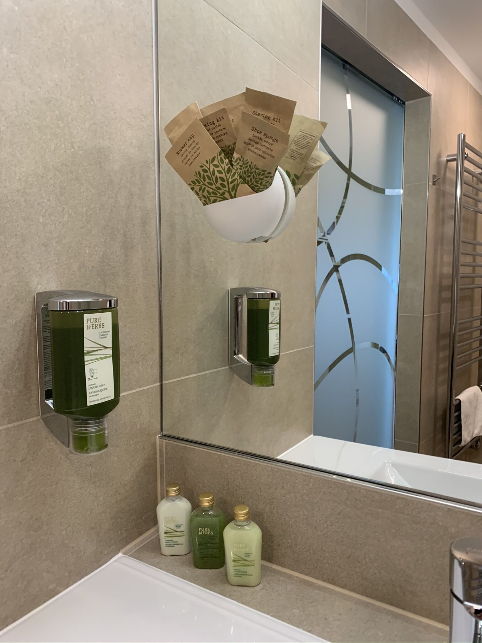 A bathroom with a large mirror, a shelf with three bottles of Pure Herbs liquid soap, and a hanging towel rack.
