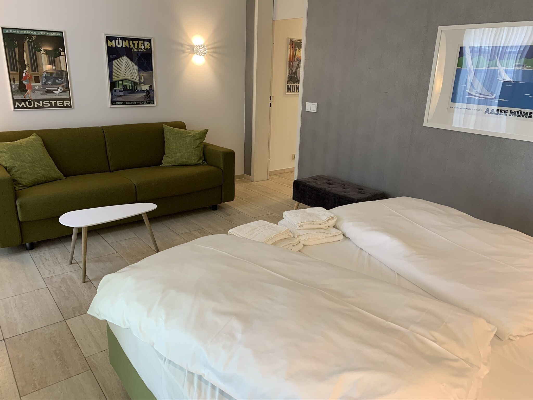 A hotel room with a bed, a green sofa, a small white table, and wall art depicting cityscapes.