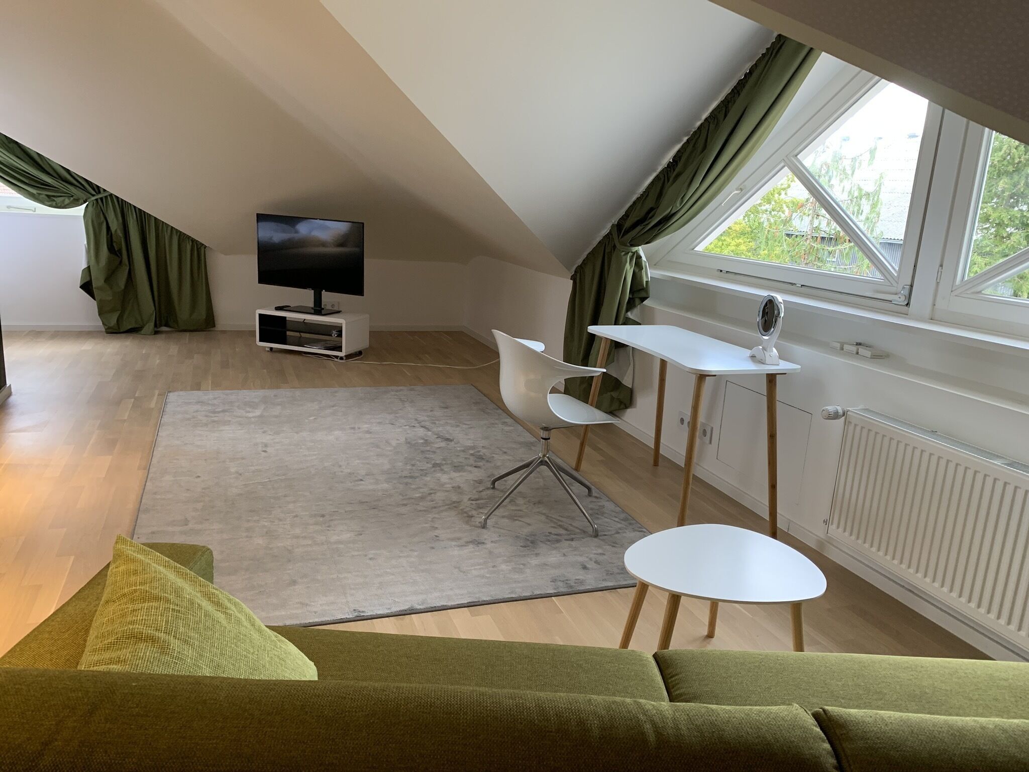 A modern loft-style room with a high ceiling, a large window with curtains, a flat-screen TV on a white stand, a green sofa, a small dining table with chairs, and a radiator.