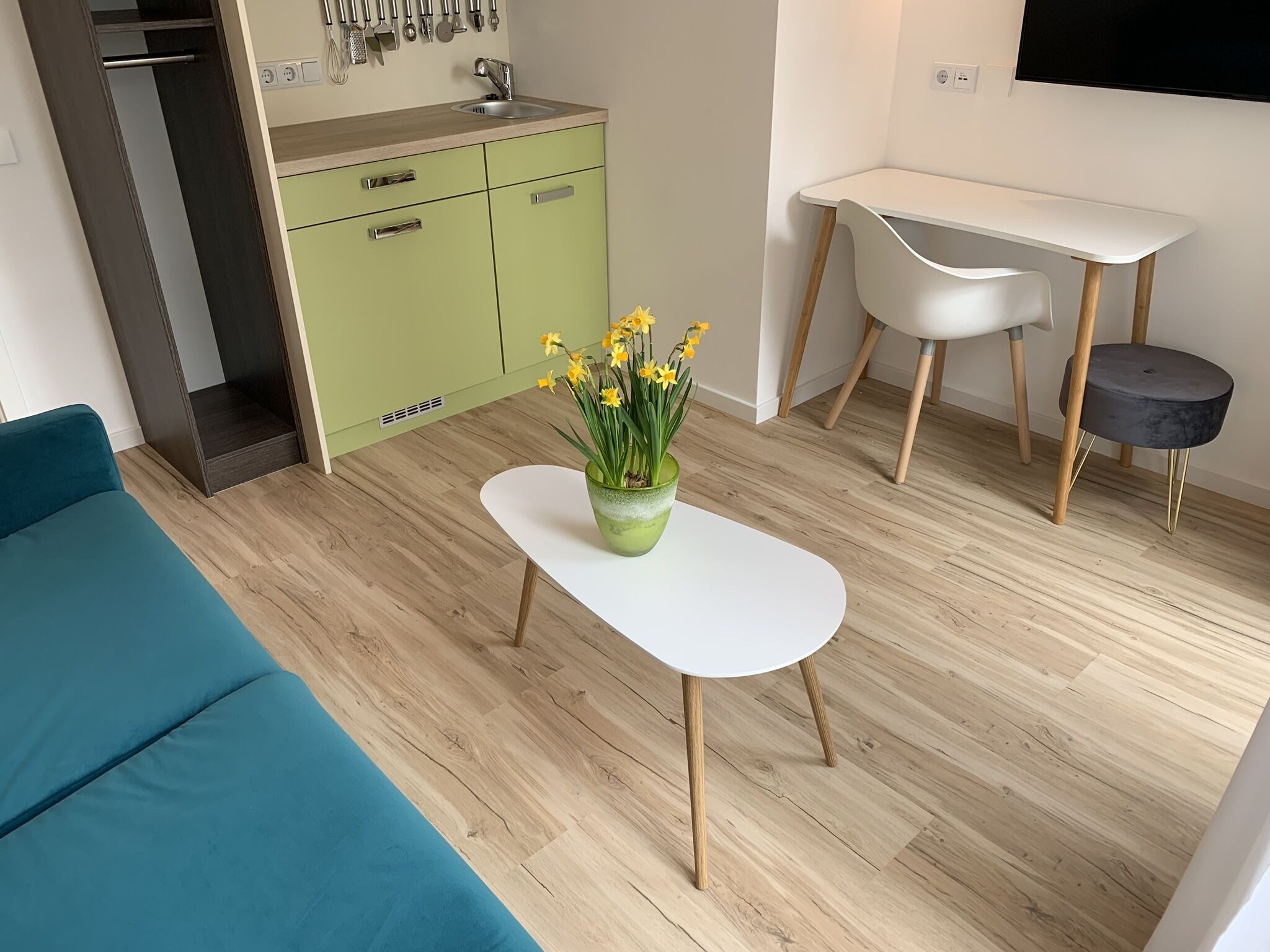 A modern kitchen with green cabinets, a white countertop, and a sink. There is a white dining table with a chair and a small stool. A vase with yellow flowers is on a white table.