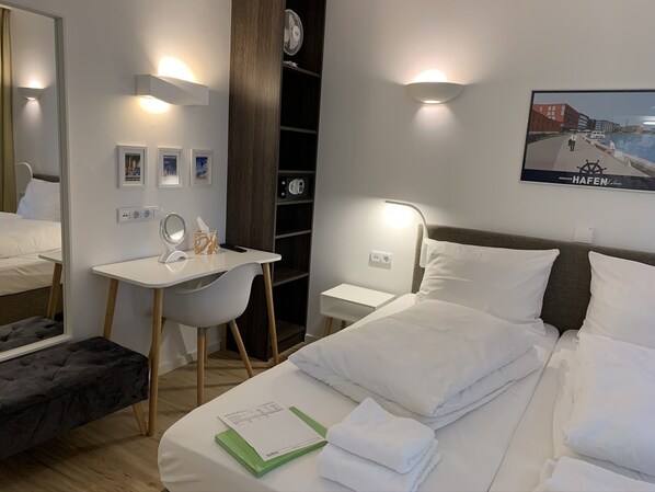 Habitación de hotel con cama, mesitas de noche, escritorio, silla, espejo, ventilador de pared y un cuadro enmarcado.