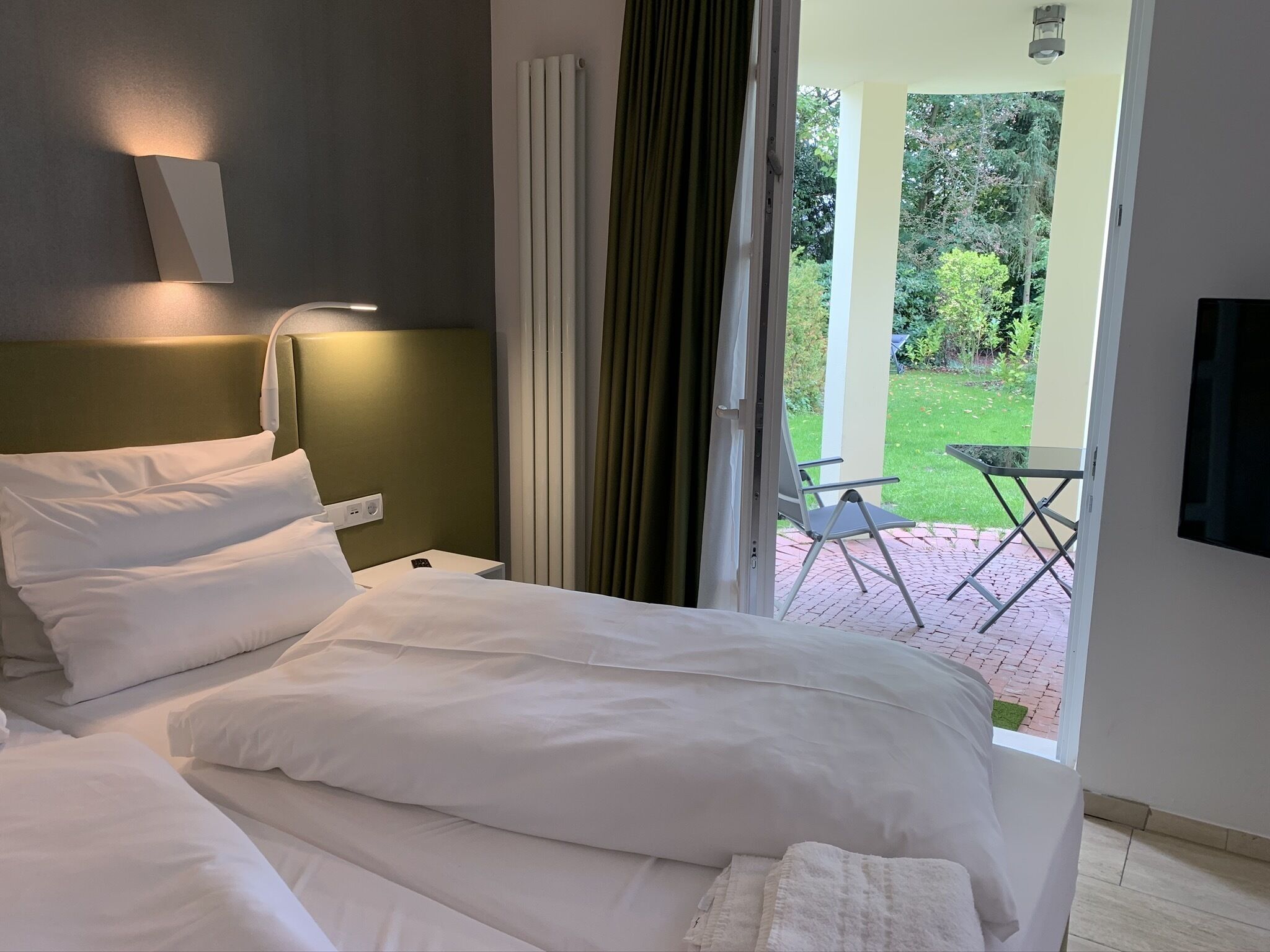 A hotel room with a bed, a bedside table with a lamp, and a view of a garden through an open door.